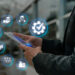 A person in a suit holds a smartphone, interacting with digital icons floating above it. Icons include a checklist, person, package, truck, warehouse, gears, and factory—symbolizing BPA in logistics and management concepts within a blurred outdoor setting. | LESSINVEST