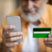 A bearded individual in an orange shirt holds a smartphone and a green credit card, showcasing the convenience of fully digital card issuance via mobile. The background remains blurred, highlighting the seamless interaction between device and card. | LESSINVEST