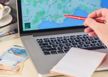 A hand with a red pen points at a laptop screen displaying a map, hinting at cross-border taxation strategies. In front of the laptop, a blank notepad awaits ideas, while euro banknotes and a steaming cup of coffee sit on the desk next to it. | LESSINVEST