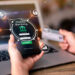 A person holds a smartphone showing a mobile banking app's login screen, symbolizing digital banking in modern investment strategies. Icons for money, security, and connectivity encircle it. Another hand grips a credit card while an open laptop hums quietly in the background. | LESSINVEST