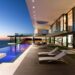 Modern luxury villa in Cyprus with a sleek design featuring floor-to-ceiling glass windows. An infinity pool stretches along the deck with several lounge chairs, all set against a serene ocean view at sunset. | LESSINVEST
