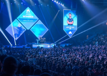 A large, packed arena hosts a gaming industry tournament on stage. Two giant screens display a player's image under the word "Winner." The audience watches intently in the dimly lit venue, with blue lights casting an electric glow over the scene. | LESSINVEST