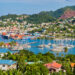 A scenic view of Grenada Island's harbor features numerous sailboats docked on the serene waters. The shoreline is adorned with colorful buildings and lush vegetation, while hills rise in the background, blanketed with greenery and scattered homes. | LESSINVEST