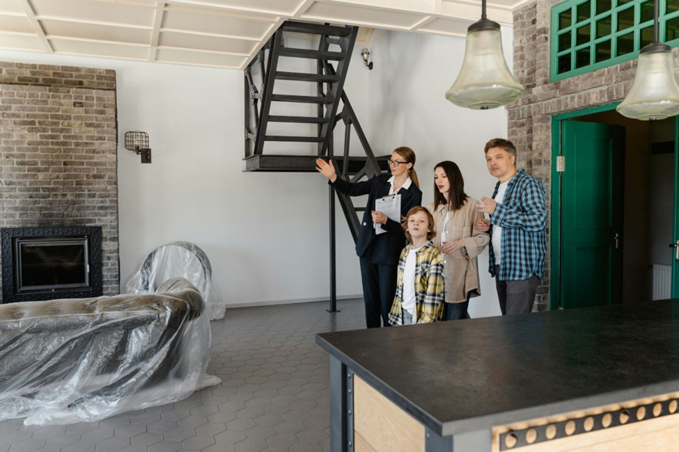 A family is being shown a room by a real estate agent. The room has a modern interior with brick walls, a black metal staircase, a fireplace, and furniture covered in plastic. The family looks engaged and interested. | LESSINVEST
