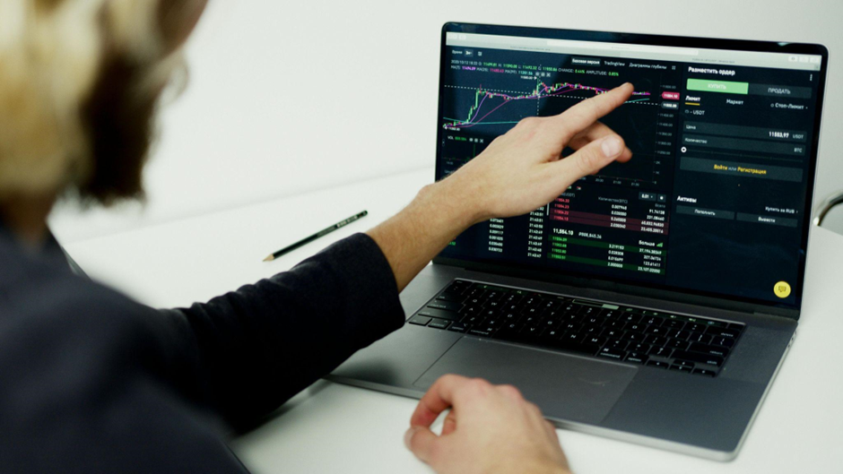 Person pointing at a laptop screen displaying stock market data, including charts and figures. A pencil is placed on the white desk beside the laptop. | LESSINVEST
