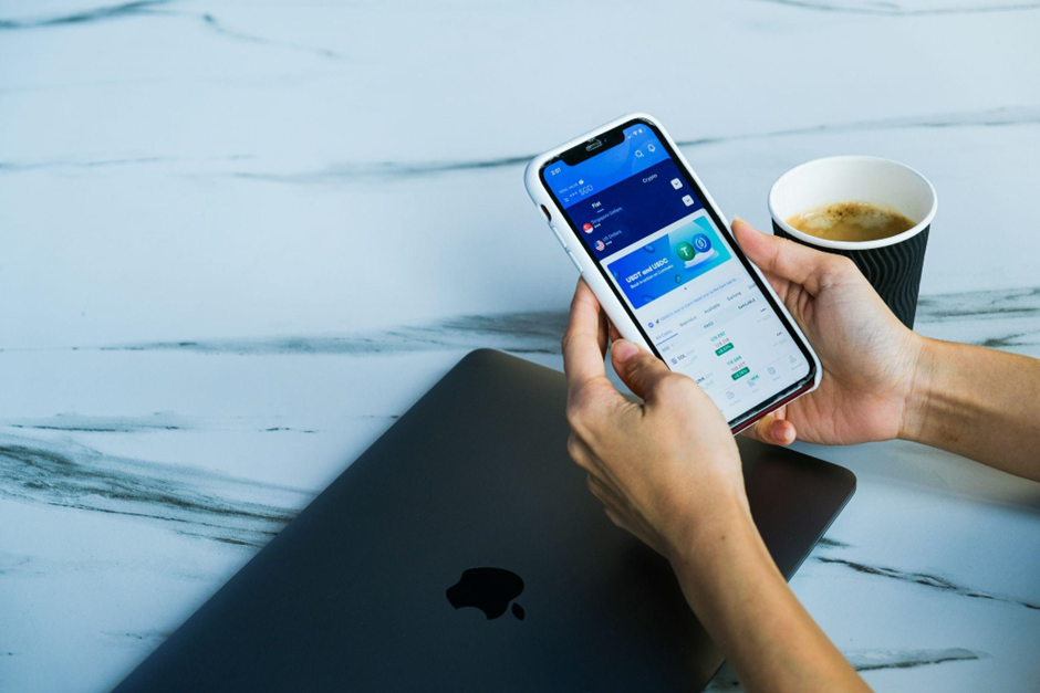 Person holding a smartphone displaying an app with data charts, next to a laptop and a cup of coffee on a marble table. | LESSINVEST