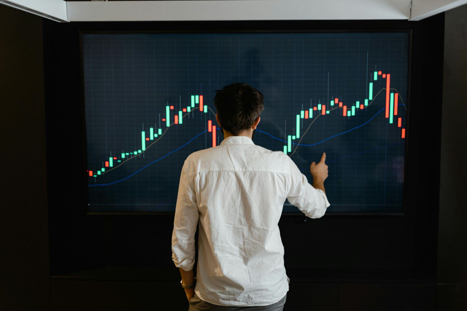 Person in a white shirt analyzing a financial chart displayed on a large screen. The chart shows colorful candlestick patterns indicating market trends against a dark background. The person points at the screen, suggesting engagement in data analysis. | LESSINVEST