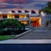 Modern building with a concrete facade and large glass windows, warmly illuminated in the evening—a gem in Toronto's real estate market. A wide staircase leads to the entrance, while surrounding trees and a vibrant sunset sky enhance the serene atmosphere. | LESSINVEST
