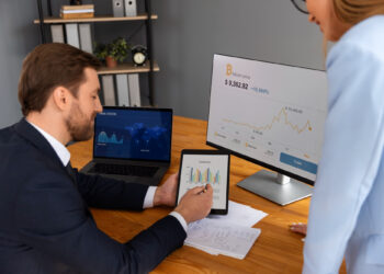A man in a suit shows a tablet with bar charts to a woman in an office, streamlining financial operations. A monitor displays Bitcoin price charts, while a laptop shows a world map. Papers and documents are on the wooden desk. | LESSINVEST