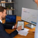 A man in a suit shows a tablet with bar charts to a woman in an office, streamlining financial operations. A monitor displays Bitcoin price charts, while a laptop shows a world map. Papers and documents are on the wooden desk. | LESSINVEST