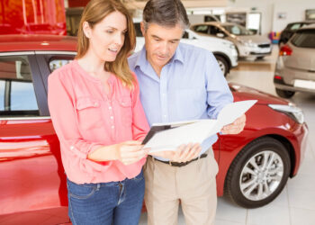 A woman and a man stand together in a car showroom, buying a used car. They're reviewing documents next to a red vehicle, with various other cars visible in the background. The woman wears a pink blouse, while the man sports a blue shirt. | LESSINVEST