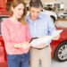 A woman and a man stand together in a car showroom, buying a used car. They're reviewing documents next to a red vehicle, with various other cars visible in the background. The woman wears a pink blouse, while the man sports a blue shirt. | LESSINVEST