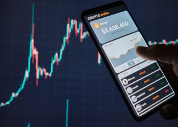 A person holds a smartphone showing a cryptocurrency wallet app with a Bitcoin balance of $5,638.450 and a graph. In the background, a larger screen displays a fluctuating line graph, indicating dynamic market trends in Cryptocurrency Trading values. | LESSINVEST