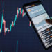 A person holds a smartphone showing a cryptocurrency wallet app with a Bitcoin balance of $5,638.450 and a graph. In the background, a larger screen displays a fluctuating line graph, indicating dynamic market trends in Cryptocurrency Trading values. | LESSINVEST