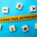 A yellow strip labeled "LONG TAIL KEYWORDS" is surrounded by six Scrabble letter tiles on a blue background. | LESSINVEST