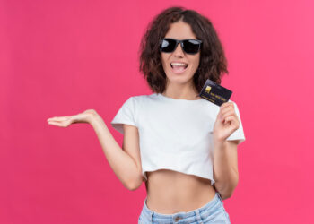 A woman with curly hair, wearing sunglasses and a white crop top, smiles as she holds a credit card—part of America’s Top Credit Cards for 2025—in one hand, gesturing confidently against a pink background. Unlock big rewards and elevate your style with every swipe. | LESSINVEST