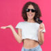 A woman with curly hair, wearing sunglasses and a white crop top, smiles as she holds a credit card—part of America’s Top Credit Cards for 2025—in one hand, gesturing confidently against a pink background. Unlock big rewards and elevate your style with every swipe. | LESSINVEST