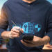 A person wearing a dark shirt uses a smartphone and stylus, exploring VPNs. Holographic icons of a keyhole, globe, and shield float above the phone, symbolizing internet security. The city skyline is blurred in the background. | LESSINVEST