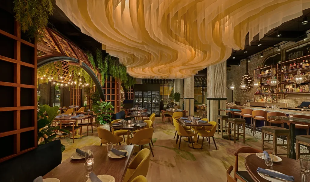 Alla Vita restaurant with round tables, yellow chairs, greenery, and a dramatic wavy ceiling installation. Warm lighting and a stylish bar create a cozy, upscale atmosphere. | LESSINVEST