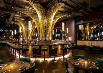 Aqua NYC in New York City restaurant with dramatic gold, tree-like light fixtures, a central bar surrounded by high chairs, round dining tables with green velvet chairs, and large mirrors on dark walls. | LESSINVEST