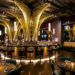 Aqua NYC in New York City restaurant with dramatic gold, tree-like light fixtures, a central bar surrounded by high chairs, round dining tables with green velvet chairs, and large mirrors on dark walls. | LESSINVEST