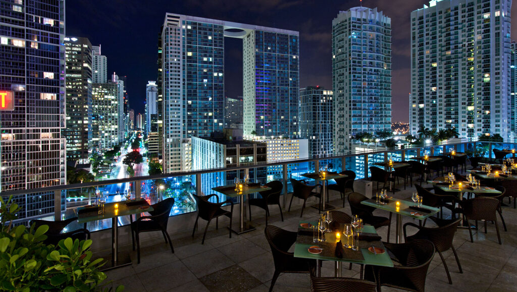 Area 31 - Downtown Miami Restaurant | Kimpton EPIC Hotel restaurant with candlelit tables overlooks a vibrant cityscape of tall, illuminated skyscrapers at night. The atmosphere is elegant, with modern buildings and city lights creating a lively backdrop. | LESSINVEST