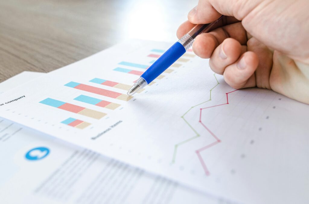 A hand holding a blue pen points to a bar chart and a line graph printed on paper, analyzing business data on a wooden desk. | LESSINVEST