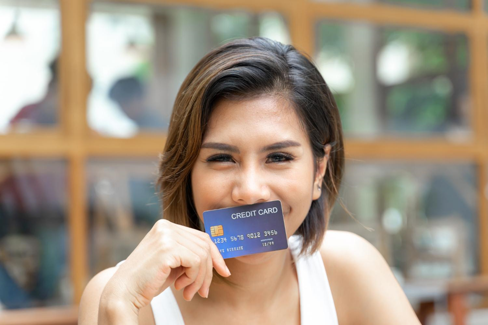 A woman with short brown hair smiles while holding a credit card in front of her face. She wears a white sleeveless top. The background is slightly blurred, showing wooden frames and greenery. | LESSINVEST