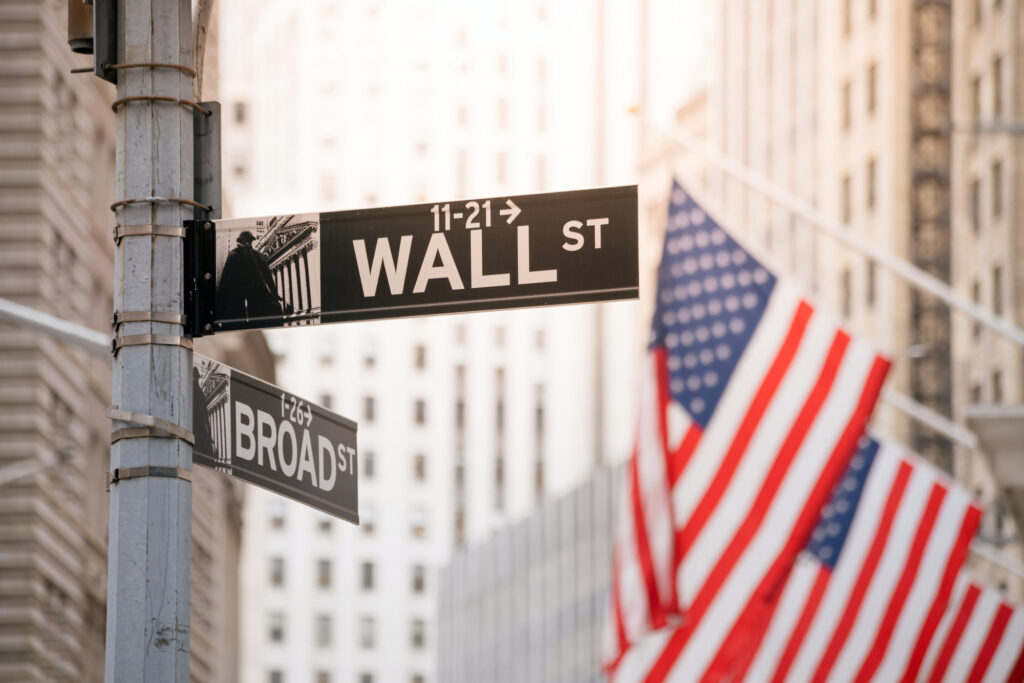 Street signs for Wall St and Broad St in New York City, surrounded by tall buildings and several American flags, highlight a hub where traditional finance meets the emerging world of Web3. | LESSINVEST