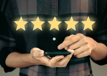 A person holding a smartphone with one hand and using the other to tap the screen. Five large yellow stars appear above the phone, representing a five-star peer review or rating. | LESSINVEST
