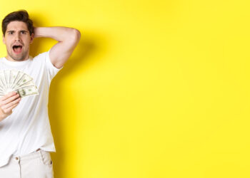 A surprised man in a white t-shirt holds a fan of U.S. dollar bills in one hand, touching the back of his head with the other, embodying the thrill of Gambling as a Hobby: How to Enjoy It Without Wrecking Your Finances, against a bright yellow background. | LESSINVEST