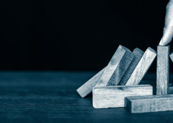 A hand uses one finger to stop a row of wooden domino blocks from falling, with several blocks already toppled on a dark wooden surface against a black background. | LESSINVEST