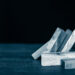 A hand uses one finger to stop a row of wooden domino blocks from falling, with several blocks already toppled on a dark wooden surface against a black background. | LESSINVEST