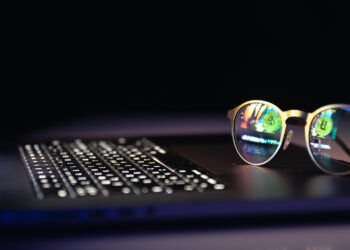 A pair of eyeglasses rests on a laptop keyboard. Computer code and a green Web3 symbol are reflected in the lenses, suggesting programming or cybersecurity work. The background is dark and out of focus. | LESSINVEST