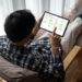 A person in a plaid shirt reclines in a chair, using a tablet displaying a workflow or project management app with colored rows and columns. Soft daylight comes through sheer curtains in the background. | LESSINVEST