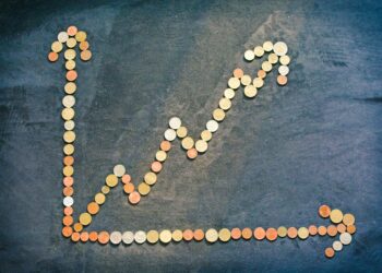A line graph made of assorted coins on a dark surface shows a rising trend, with two arrows—one pointing upward and one to the right—representing financial or business growth. | LESSINVEST