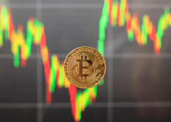 A gold Bitcoin coin is centered in front of a blurred background showing a colorful, fluctuating Bitcoin Price Live chart with red and green candlestick lines. | LESSINVEST