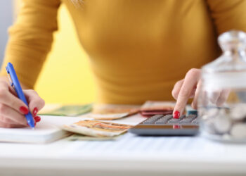 A person in a yellow top uses a calculator with one hand and writes on a notepad with the other, creating a budget. Euro banknotes and a jar of coins are on the table. | LESSINVEST