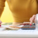 A person in a yellow top uses a calculator with one hand and writes on a notepad with the other, creating a budget. Euro banknotes and a jar of coins are on the table. | LESSINVEST