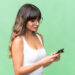 A woman with long brown hair and glasses, wearing a white tank top, is looking at and using her smartphone—possibly exploring the Hint App—against a plain green background. | LESSINVEST