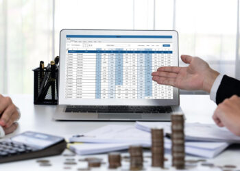 Two people in business attire sit at a desk with coins, documents, and a calculator. A laptop between them displays financial data as one gestures toward the screen, discussing Balance Sheet Reconciliation. | LESSINVEST