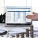 Two people in business attire sit at a desk with coins, documents, and a calculator. A laptop between them displays financial data as one gestures toward the screen, discussing Balance Sheet Reconciliation. | LESSINVEST