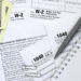 A close-up of tax documents, including 1040 and W-2 forms, with a pen, a notepad, paper clips, and several $100 bills scattered on a desk. | LESSINVEST