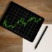 A tablet displaying a green fluctuating line graph rests on a wooden desk next to a blank sheet of paper and a black pen. | LESSINVEST