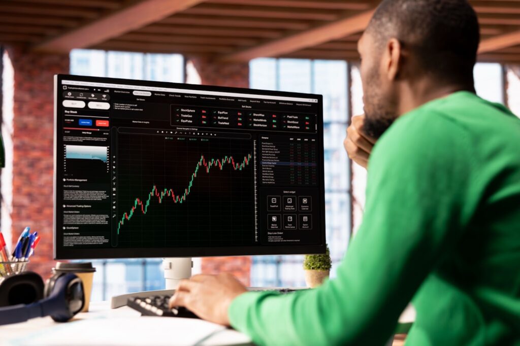 A person in a green shirt looks at a large computer monitor displaying a financial trading platform with charts, QSBS insights, and data in a bright, modern office space. | LESSINVEST