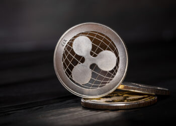 A close-up of a Ripple (XRP) cryptocurrency coin standing upright on two other coins, set against a dark, blurred background, symbolizes how Ripple XRP shifts to Swift-like solutions in global payments. | LESSINVEST