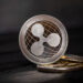 A close-up of a Ripple (XRP) cryptocurrency coin standing upright on two other coins, set against a dark, blurred background, symbolizes how Ripple XRP shifts to Swift-like solutions in global payments. | LESSINVEST