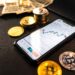 A smartphone displaying a cryptocurrency trading chart is placed on a dark table, surrounded by gold Bitcoin coins, a silver Litecoin coin, and stacks of US dollar bills and coins in the background. | LESSINVEST