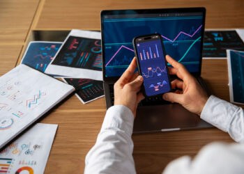 A person holds a smartphone displaying financial graphs next to an open laptop with charts, surrounded by printed reports—illustrating the integration of digital assets with traditional investments on a wooden desk. | LESSINVEST