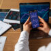 A person holds a smartphone displaying financial graphs next to an open laptop with charts, surrounded by printed reports—illustrating the integration of digital assets with traditional investments on a wooden desk. | LESSINVEST